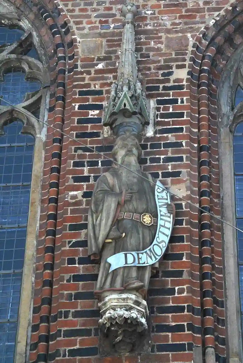 Demosthenes - ursprünglich Prophet Jesaja - trägt um sein Gewand den Dupfing, unter dem Gewand bildet sich eine Tasche ab. Seine linke Hand umfasst sein Spruchband, die Rechte liegt auf der Brust. Da der ursprüngliche Kopf dieser Figur nicht erhalten geblieben ist, musste er ersetzt werden.