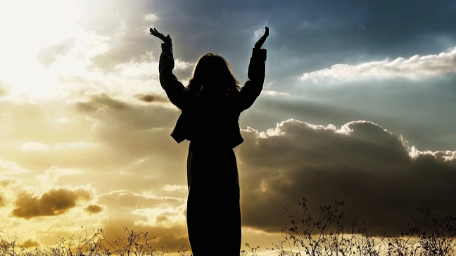 Symbolbild für Religionsangelegenheiten. Eine Frau steht auf einer Wiese und hält ihre Hände Richtung Himmel hoch.